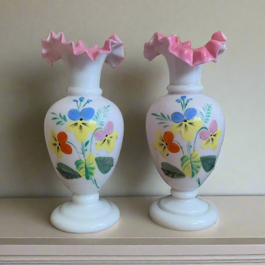 A pair of white glass vases with crimped ruffle rims. They have hand painted pansies on the front of each vase.