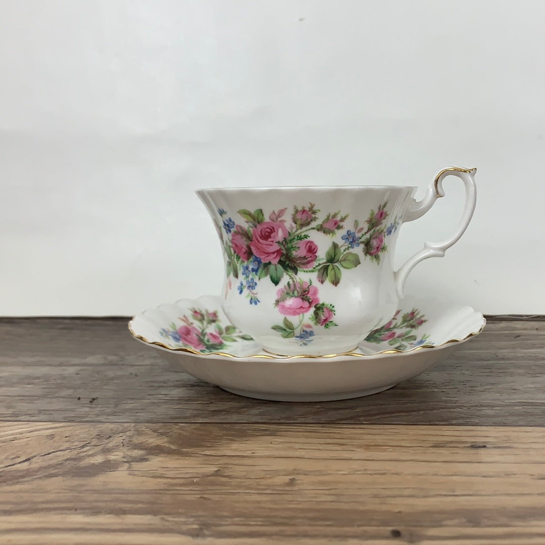 A white ceramic teacup with a floral pattern, featuring pink and blue roses, on a matching saucer with gold trim.