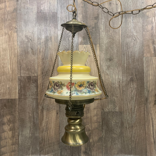 A vintage swag lamp with a cased glass floral shade in yellow and white, with a brass-colored metal base and frame, hanging against a wooden background.